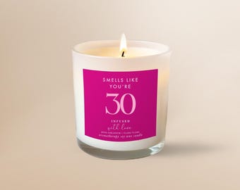 Może przedstawiać: Biała świeca w matowym szkle z zapalonym knotem i jaskraworóżową etykietą. Etykieta zawiera napis "SMELLS LIKE YOU'RE 30", "INFUSED WITH LOVE" oraz "ROSE GERANIUM + YLANG YLANG" białymi i różowymi literami na świecy z wosku sojowego.