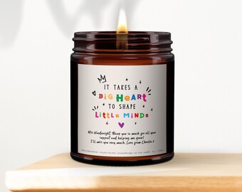 Puede incluir: Una vela encendida en un frasco de vidrio ámbar con una etiqueta blanca que presenta el texto IT TAKES A BIG HEART TO SHAPE LITTLE MINDS en letras coloridas, un mensaje personalizado para Miss Woodwright de Charlie y el texto 180ml.