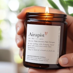Peut inclure: Une bougie d'aromathérapie allumée dans un pot en verre brun, tenue par une personne. La bougie a une étiquette blanche avec le mot "Airapist" et une définition. La bougie est faite de cire de soja et d'huiles essentielles, et est coulée à la main en Angleterre.