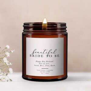 Pode incluir: Uma vela em frasco de vidro castanho com um rótulo branco apresentando o texto Beautiful BRIDE TO BE, Happy Hen Weekend! Lots of love, Sarah, Becs, Tina, Karen, e YLANG YLANG + LAVENDER soy wax candle 6oz / 180ml. A vela tem um pavio aceso.