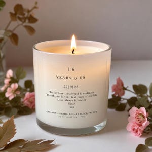 Op de afbeelding: Een brandende sojawas kaars in een doorzichtige glazen pot met een wit etiket. De tekst luidt 16 YEARS OF US, 22|9|23, een persoonlijke boodschap aan een zielsverwant en ORANGE + SANDALWOOD + BLACK PEPPER, 6oz / 180ml. De kaars geeft warm licht.