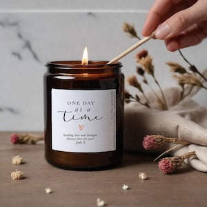 Op de afbeelding: Een brandende bruine glazen potkaars met de tekst "ONE DAY at a time" op een wit etiket. De kaars wordt aangestoken met een lucifer. Gedroogde bloemen en een linnen doek staan op de achtergrond. De geur van de kaars is citroengras, lavendel en eucalyptus.