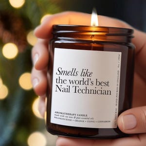 Puede incluir: Una vela de aromaterapia encendida en un tarro de vidrio marrón, sostenida por una persona. La etiqueta blanca dice "Smells like the world's best Nail Technician". También indica que está vertida a mano en Inglaterra, 180 ml, y enumera los ingredientes.