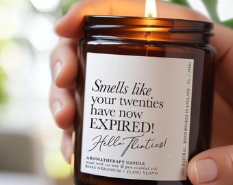Könnte beinhalten: Eine brennende Aromatherapie-Kerze in einem braunen Glasgefäß mit einem weißen Etikett, auf dem steht: Smells like your twenties have now EXPIRED! Hello Thirties! Aromatherapy candle made with soy wax and pure essential oils, Rose Geranium + Ylang Ylang, 180ml.