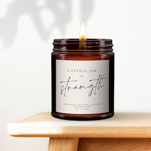Op de afbeelding: Een brandende sojawas kaars in een bruine glazen pot met een wit etiket met de tekst A LITTLE JAR of strength, Lemongrass + Lavender + Eucalyptus, 6oz / 180ml. De kaars heeft een enkele brandende lont die een warme vlam afgeeft.