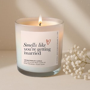 May include: A burning aromatherapy candle in a frosted glass jar. The label says "Smells like you're getting married" with a heart symbol. The candle is made with soy wax and essential oils, and is 6 oz / 180ml.