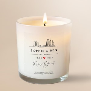 Op de afbeelding: Een witte glazen kaars met een brandende lont, voorzien van een zwarte lijntekening van de skyline van New York. Het etiket vermeldt SOPHIE & BEN ENGAGED 15.03 2025 met een rood hartje, New York, en Rose Geranium + Ylang Aromatherapy soy wax candle.