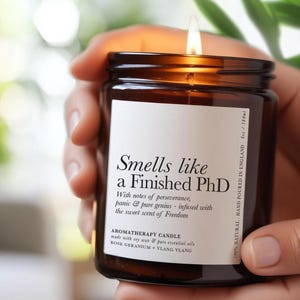 Könnte beinhalten: Eine brennende Aromatherapie-Kerze in einem braunen Glas. Das Etikett trägt den Text "Smells like a Finished PhD". Die Kerze besteht aus Sojawachs und ätherischen Ölen und hat 180 ml. Handgegossen in England.