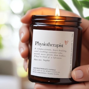 Könnte beinhalten: Eine brennende Aromatherapie-Kerze in einem braunen Glas, gehalten in einer Hand. Das Etikett lautet "Physiotherapeut" mit einer Beschreibung und "AROMATHERAPIE-KERZE" Details. Die Kerze ist 180 ml und wird aus Sojawachs und ätherischen Ölen hergestellt.