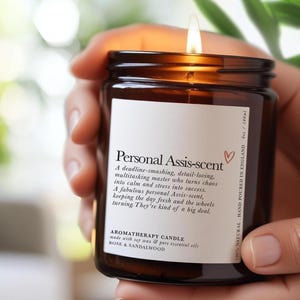 Könnte beinhalten: Eine brennende Aromatherapie-Kerze in einem braunen Glas, gehalten von einer Person. Das Etikett lautet "Personal Assis-scent" und listet die Inhaltsstoffe auf. Die Kerze hat 180 ml und wird aus Sojawachs und ätherischen Ölen hergestellt.