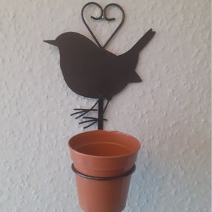 Puede incluir: Un macetero de pared con forma de pájaro de metal negro con un detalle de corazón sobre la cabeza del pájaro. El macetero contiene una pequeña maceta de color terracota. La pared es de color blanco roto texturizado.
