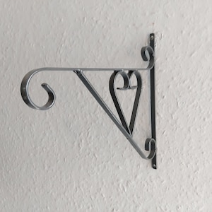Pode incluir: Suporte de prateleira de metal preto com um design em forma de coração. O suporte é fixado a uma parede com um parafuso.