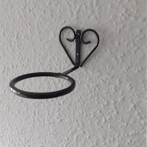 Op de afbeelding: Zwarte metalen plantenhanger met een hartvormig ontwerp en een ronde ring. De hanger is gemonteerd op een witte muur.
