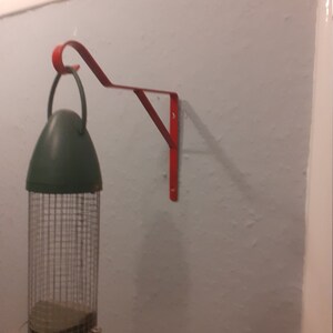 Puede incluir: Un soporte de metal rojo para comedero de pájaros montado en una pared con un comedero de pájaros verde y plateado colgando.