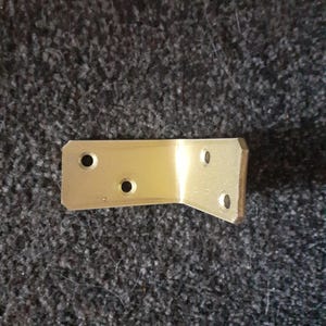 May include: A gold metal corner brace with four holes for screws. The brace is designed to reinforce corners and joints in furniture or other structures.