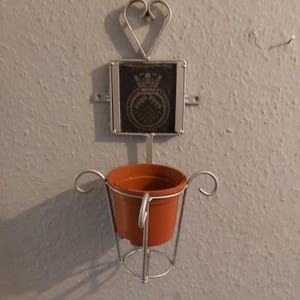 May include: A silver metal plant hanger with a heart-shaped design at the top. The hanger has a small, brown terracotta pot attached to it. The pot is empty.