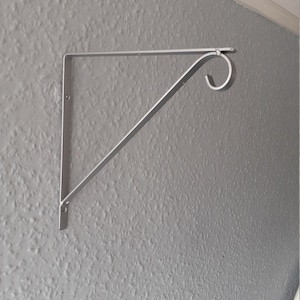 Könnte beinhalten: Eine silberne Metall-Regalhalterung mit einer dekorativen Rolle am Ende. Die Halterung ist an einer Wand montiert.
