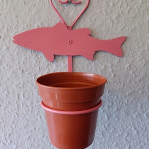 May include: A pink metal wall planter with a fish-shaped sign and a heart-shaped top. The planter holds a brown plastic pot. The fish sign is attached to the wall with a screw. The pot is held in place by a pink metal ring.