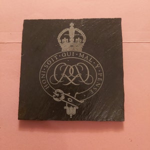 Puede incluir: Posavasos cuadrado de pizarra negra con diseño grabado. El diseño incluye una corona, letras entrelazadas y el texto "HONI SOIT QUI MAL Y PENSE". Un artículo decorativo para el hogar.