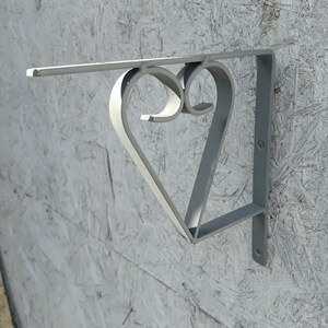 Puede incluir: Un soporte de estante de metal plateado con un diseño en forma de corazón. El soporte está montado en una pared.