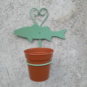 May include: A light green metal fish-shaped planter with a heart-shaped hanger. A terracotta pot is hanging from the fish's mouth.