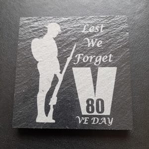 May include: A black slate coaster with a white silhouette of a soldier holding a rifle. The text "Lest We Forget" is above a white "V" with the number "80" inside. The text "VE DAY" is below the "V".