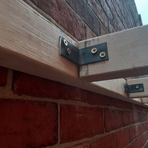 May include: Close-up of a wooden beam attached to a brick wall with metal brackets and screws. The brackets are secured with screws.