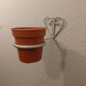 Könnte beinhalten: Ein silberner Pflanzenhalter aus Metall in Herzform mit einem Terrakotta-Topf. Der Halter ist an einer weißen Wand befestigt.
