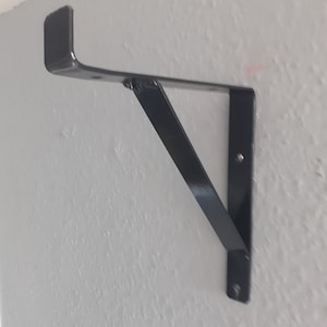 May include: Black metal shelf bracket with a straight arm and a right angle. The bracket is mounted to a white wall.