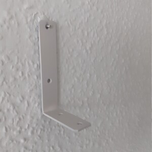 May include: White metal L-shaped bracket with three screw holes. The bracket is mounted to a white wall.