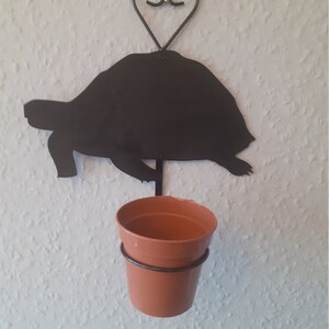 Puede incluir: Macetero de pared de silueta de tortuga de metal negro con un colgador en forma de corazón y una maceta de terracota marrón.