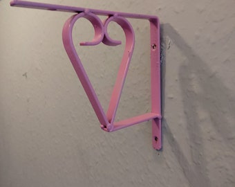 May include: A pink metal shelf bracket shaped like a heart. The bracket is mounted to a wall.