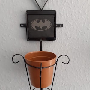 May include: Black metal wall planter with a heart-shaped top and a square frame with a black slate tile featuring the Batman logo. The planter has a brown pot.