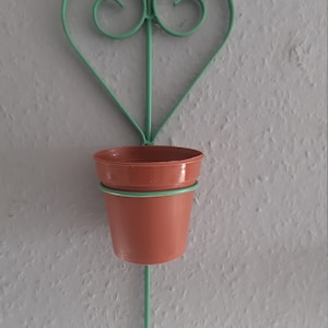 May include: A green metal heart-shaped plant hanger with a brown plastic pot attached. The hanger is designed to be mounted on a wall.