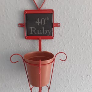 Puede incluir: Un macetero de pared de metal rojo con una parte superior en forma de corazón y una pequeña pizarra que dice "40th Ruby". La maceta tiene una maceta de color terracota y está diseñada para contener una planta pequeña. El diseño general es decorativo.