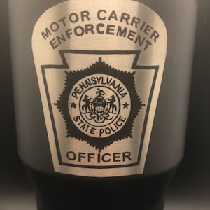 Peut inclure: Un gobelet en acier inoxydable noir avec un design gravé de couleur argent. Le design présente les mots "Motor Carrier Enforcement", un emblème de la police d'État de Pennsylvanie et le mot "Officer".