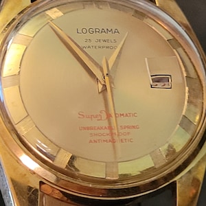 May include: A gold-toned wristwatch with a white face. The watch face has black hands and black Roman numeral hour markers. The watch face also has the text "Lograma 25 Jewels Waterproof Super Automatic Unbreakable Spring Shockproof Antimagnetic".