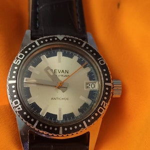 May include: A silver wristwatch with a black leather band and a white face. The watch has a black bezel with white numbers and a date window at the 3 o'clock position. The watch face also has the text "Evan 17 Rubis" and "Antichoc".
