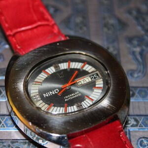 May include: A silver-toned wristwatch with a black face and red accents. The watch has a date window that displays "SAT 25". The watch face also has the text "NINO INCABLOC AUTOMATIC". The watch is on a red leather band.
