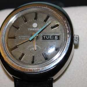 May include: A silver-toned wristwatch with a round face and a black leather band. The watch face has a silver background with black markings and a date window that reads "TUE 5". The watch also has the words "Tressa Automatic 21 Jewels Laser Beam" printed on the face.