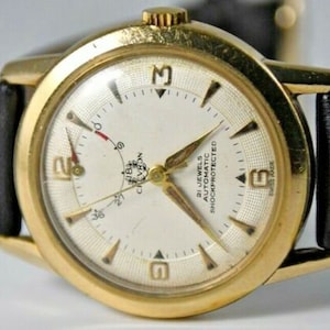 May include: A gold-tone wristwatch with a white face and black leather strap. The watch face has black Roman numeral hour markers, black hands, and the text "21 Jewels Automatic Shockprotected" printed on it.