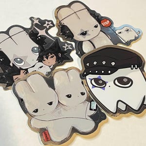 May include: A collection of die-cut stickers featuring cartoon characters with a gothic aesthetic. The stickers include a ghost wearing a black hat, a bunny with a stop sign, and other characters with a sad expression.