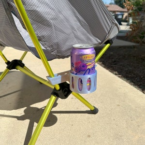 May include: A gray and yellow camping chair with a 3D printed gray cup holder attached to one of the legs. A purple can of Black Raspberry Sparkling Essence is in the cup holder.