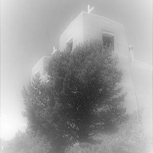 Puede incluir: Una fotografía en blanco y negro de un edificio blanco de adobe con una cruz en la parte superior. El edificio está parcialmente oscurecido por un gran árbol en primer plano.