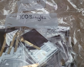 Op de afbeelding: Een doorzichtige plastic zak met verschillende kleine gouden metalen voorwerpen. De zak is gelabeld met "Inhoud: 100 Singles".