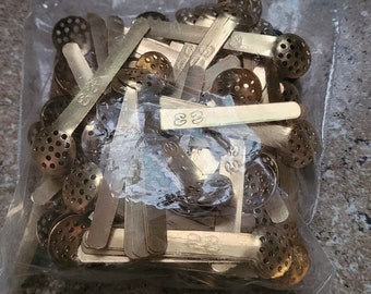 Op de afbeelding: Een doorzichtige plastic zak gevuld met een grote hoeveelheid goudkleurige metalen gaasnetten en kleine, platte, rechthoekige stukjes metaal. De metalen stukjes hebben het nummer "33" erop gestempeld.