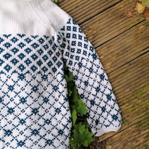 Może przedstawiać: Biały sweter dzianinowy z niebiesko-białym geometrycznym wzorem. Sweter ma długie rękawy i okrągły dekolt.