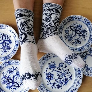May include: White knitted socks with a navy blue floral pattern are shown with several blue and white porcelain plates. The plates have a floral design and a scalloped edge. The socks are on a wooden surface.