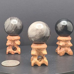 May include: Three polished gemstone spheres, each resting on a carved wooden stand. The spheres vary in color, with two displaying a mix of gray and black patterns, and one appearing solid black. A silver coin is in the foreground.