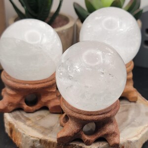 May include: Three white crystal spheres on wooden stands. The spheres are smooth and translucent, with a slight cloudy appearance. The stands are a dark brown color and have a simple design.